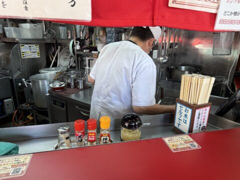 こいけのカウンター越しの厨房。亭主が調理中、手前には七味・胡椒・辣油・ニンニク油の調味料と「お水はセルフです」の札