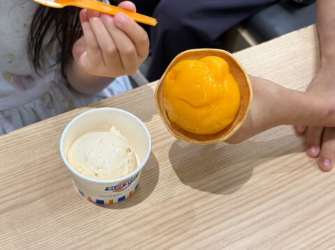 テーブルに並んだマンゴー色のコーンとカップのアイス。娘がスプーンを持つ