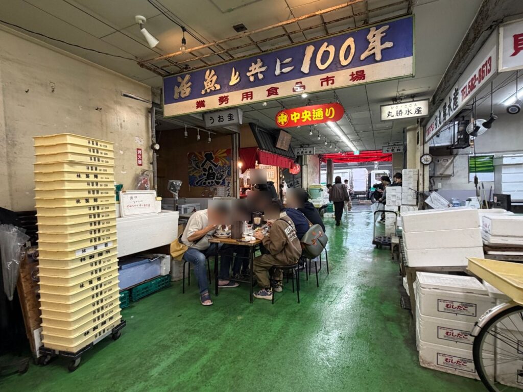 マルナカ食品センターの「活魚と共に100年」の看板と、テーブル席で朝ラーを食べる人たち